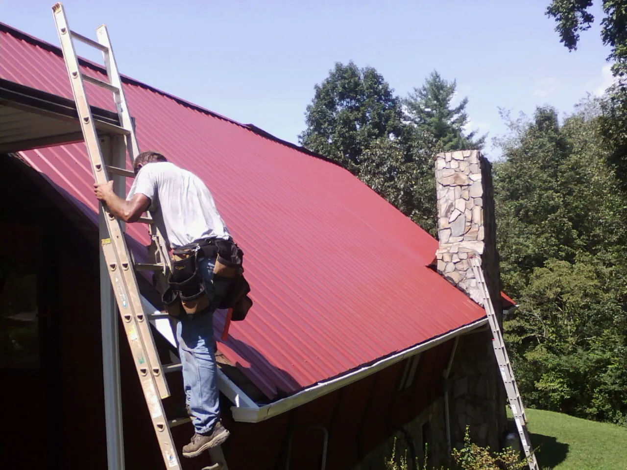 Roofer on a ladder beside a red metal roof and stone chimney — Soffit & Fascia Repair by Roofing Brothers in Arapahoe